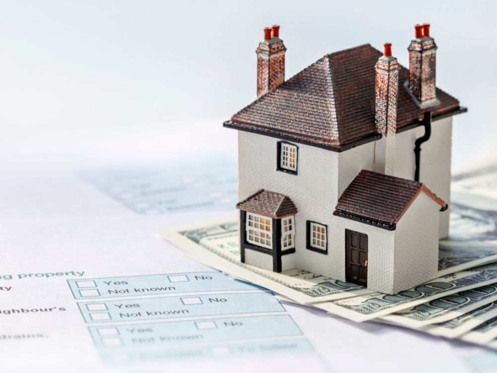 Miniatura de casa sobre dinheiro e documentos imobiliários, representando a economia obtida com o desconto cartório primeiro imóvel na compra de uma residência.